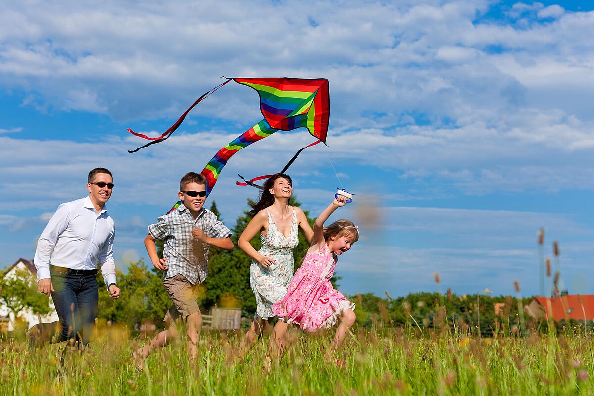 Eine Familie mit vier Personen läuft durch ein Feld und fliegt einen bunten Regenbogendrachen. Sie sind in legerer Sommerkleidung gekleidet und zeigen Freude, während sie gemeinsam unter einem teilweise bewölkten Himmel spielen.