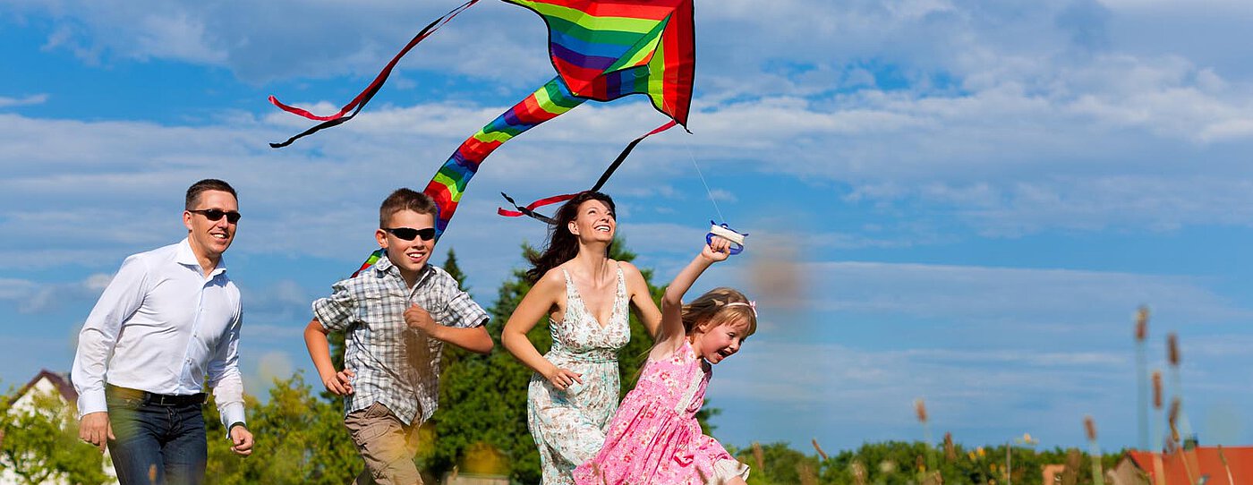 Eine Familie mit vier Personen läuft durch ein Feld und fliegt einen bunten Regenbogendrachen. Sie sind in legerer Sommerkleidung gekleidet und zeigen Freude, während sie gemeinsam unter einem teilweise bewölkten Himmel spielen.