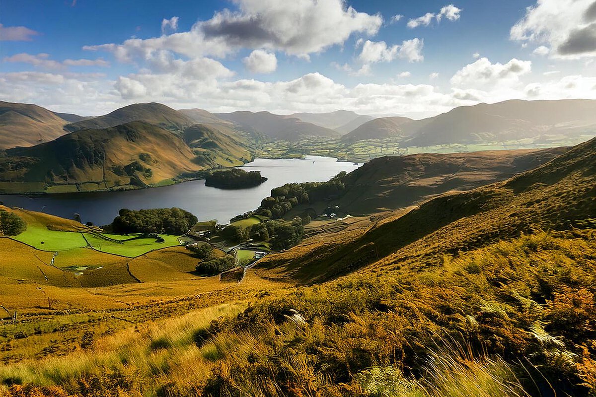 Beeindruckende Landschaft des Lake District mit sanften Hügeln, einem friedlichen See und lebhaft grünen Tälern, alles unter einem teilweise bewölkten Himmel.