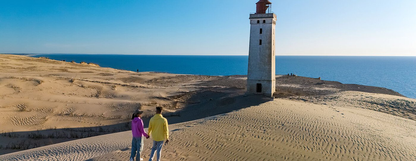 A couple holding hands stands on soft sand dunes overlooking an old lighthouse and the wide blue Danish coastline. Morning sunlight bathes the scene in clear light as the calm sea glimmers on the horizon.