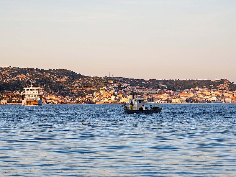 A small fishing boat and a ferry sail across calm blue waters toward a sunlit coastal town surrounded by hills and islands.