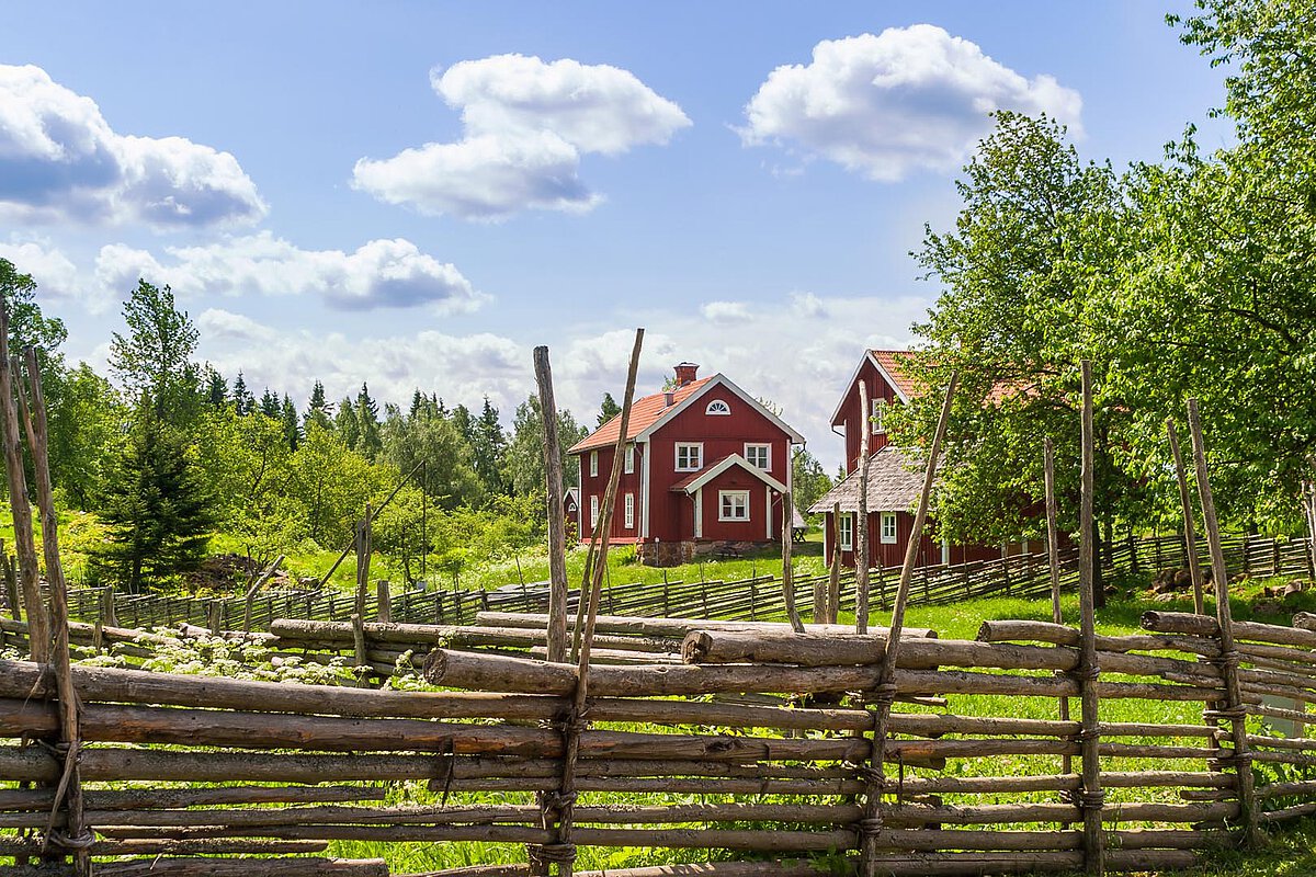 Traditionelle rote Gebäude auf einer Farm in Mittelschweden, umgeben von Grün und einem rustikalen Holzzaun, unter einem blauen Himmel mit fluffigen Wolken, die eine friedliche ländliche Umgebung darstellen.