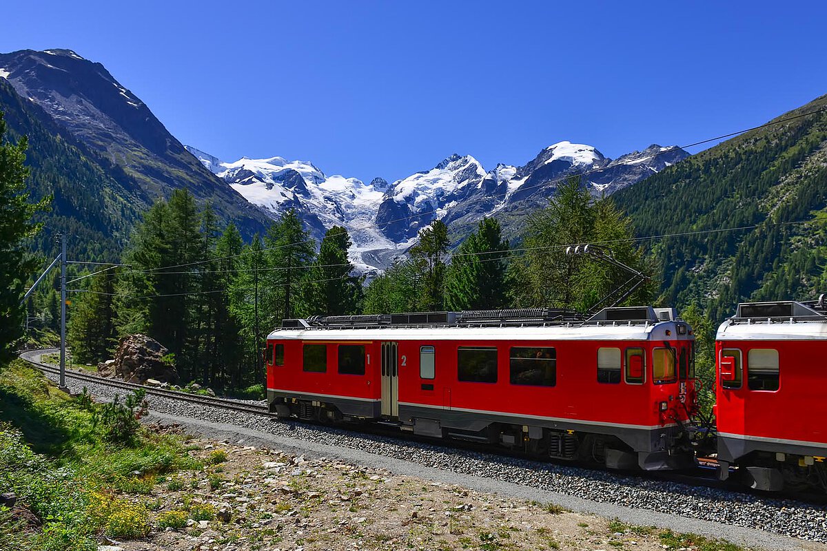 Ein roter Zug auf Gleisen in den Berner Alpen, mit schneebedeckten Bergen und Grün im Hintergrund unter einem klaren blauen Himmel.