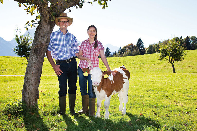 Landwirtschaftliche Unternehmer Stefanie und Stefan Boos Stefanie und Stefan Boos, landwirtschaftliche Unternehmer und Gründer von bauernhofurlaub.de und farmtravel.com, lächeln neben einem Kalb auf einem grünen Feld, mit Bäumen und Bergen im Hintergrund, während sie Arbeitskleidung und Stiefel tragen.