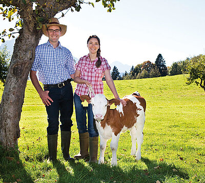 Stefanie and Stefan Boos, agricultural entrepreneurs and forunders of bauernhofurlaub.de and farmtravel.com, smile next to a calf in a green field, with trees and mountains in the background, wearing work attire and boots.