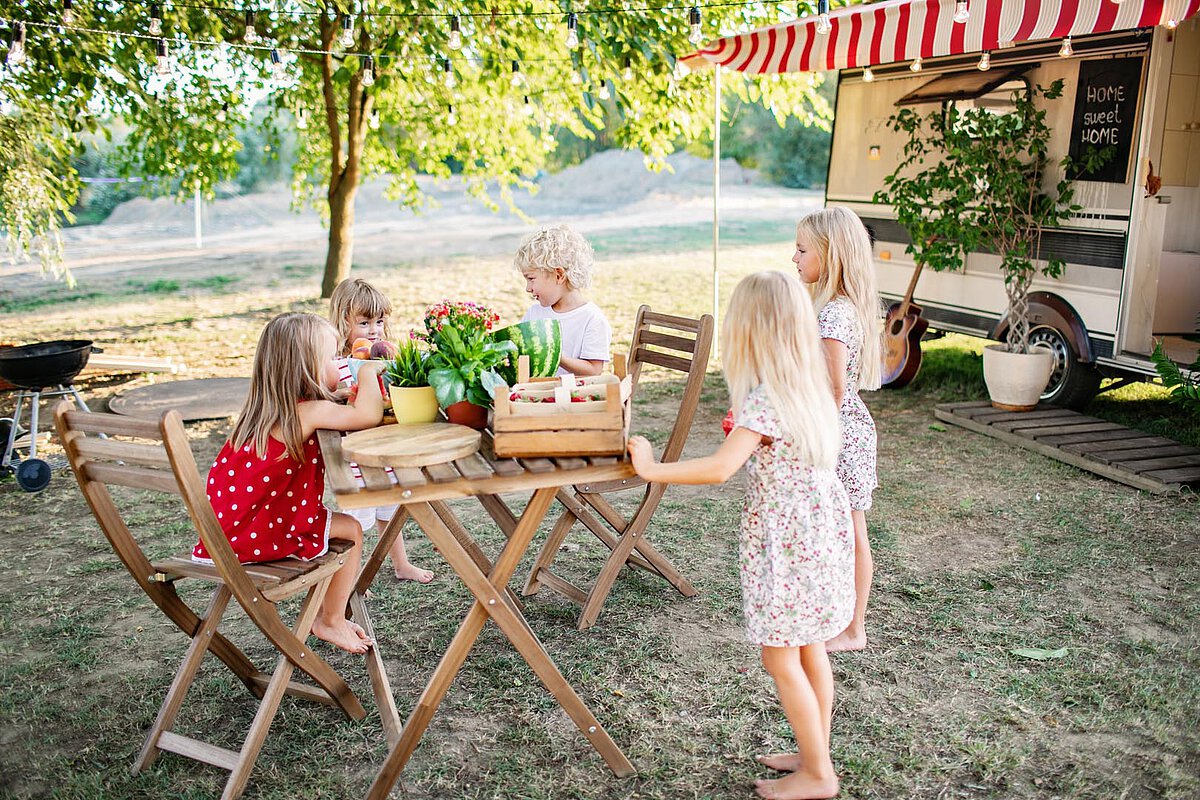 Five children sitting around a wooden table with plants and flowers, enjoying a sunny day outdoors near a trailer, surrounded by greenery.