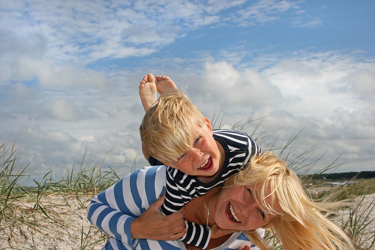 A mother carrying her son on her back at the beach, both smiling happily. They are surrounded by grass and a cloudy sky.