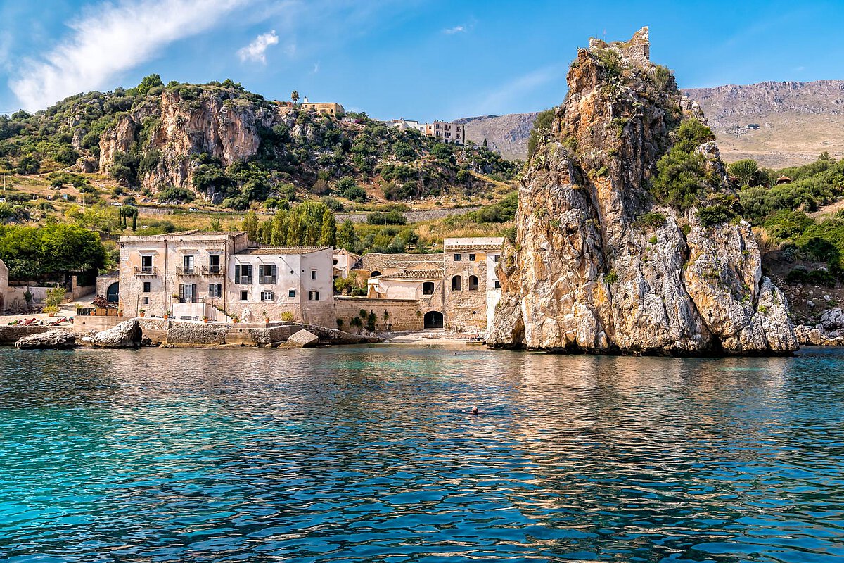 A coastal view of Scopello with historic stone buildings by the water, a rocky outcrop rising from the sea, and lush hills in the background under a clear blue sky.