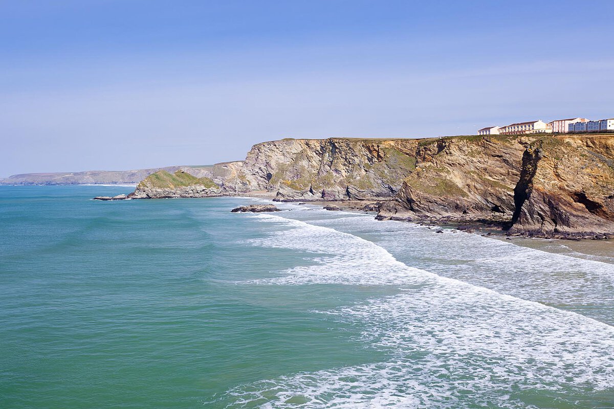 Malerischer Blick auf die Küste von Cornwall mit rauen Klippen und Wellen, die sanft am Strand plätschern, vor einem klaren blauen Himmel.