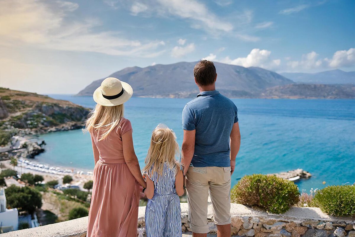 A family of three stands on a stone terrace overlooking a blue sea and mountains. The mother wears a hat and a pink dress, the daughter holds her hand in a striped dress, and the father stands beside them in a blue shirt, all enjoying the scenic view.