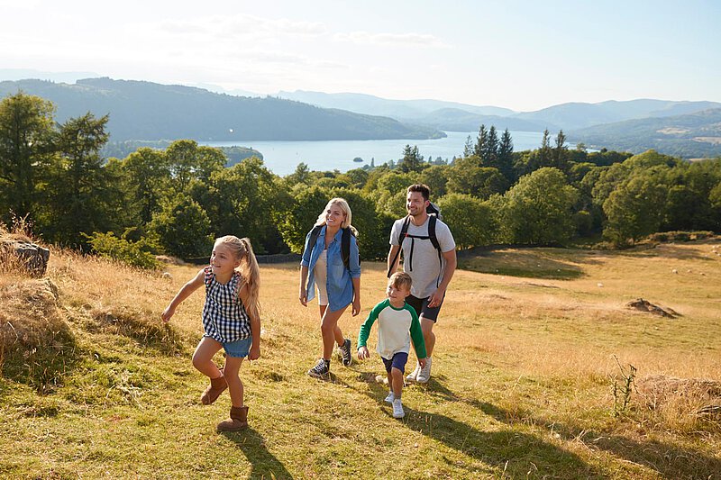 Familienwanderung mit Aussicht Eine Familie mit vier Personen wandert auf einem grünen Hang mit Blick auf einen See und Berge. Das junge Mädchen läuft voraus, während ihr Bruder hinterherläuft. Die Eltern gehen zusammen, tragen Rucksäcke und lächeln, mit einem strahlend blauen Himmel und Bäumen, die sie umgeben.