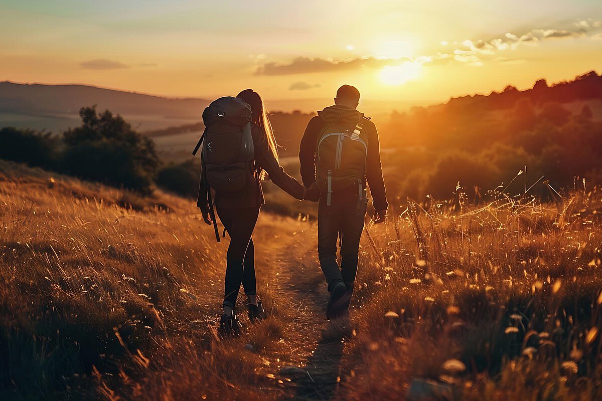 Ein Mann und eine Frau halten sich beim Wandern auf einem Pfad am Sonnenuntergang die Hände. Sie haben Rucksäcke dabei, und die Landschaft zeigt goldene Gräser und sanfte Hügel, beleuchtet vom warmen Schein der untergehenden Sonne.