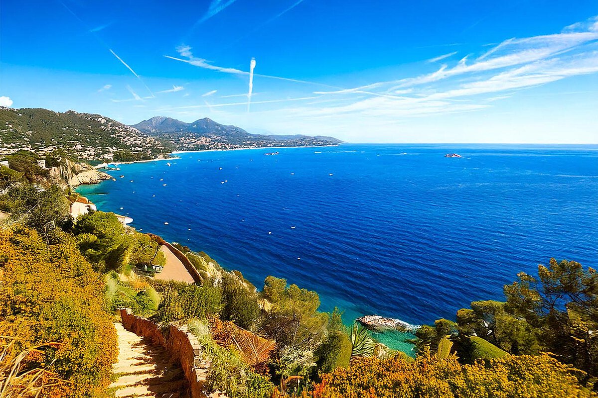 Vibrant blue waters of the Côte d'Azur with lush greenery and boats, framed by distant mountains under a clear sky.