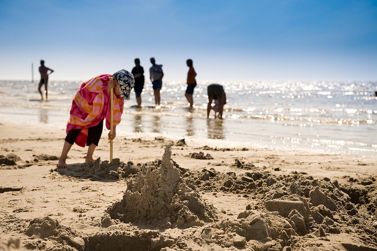 A child wearing a colorful towel kneels in the sand, building a sandcastle, while people are seen enjoying the water in the background under a bright blue sky.