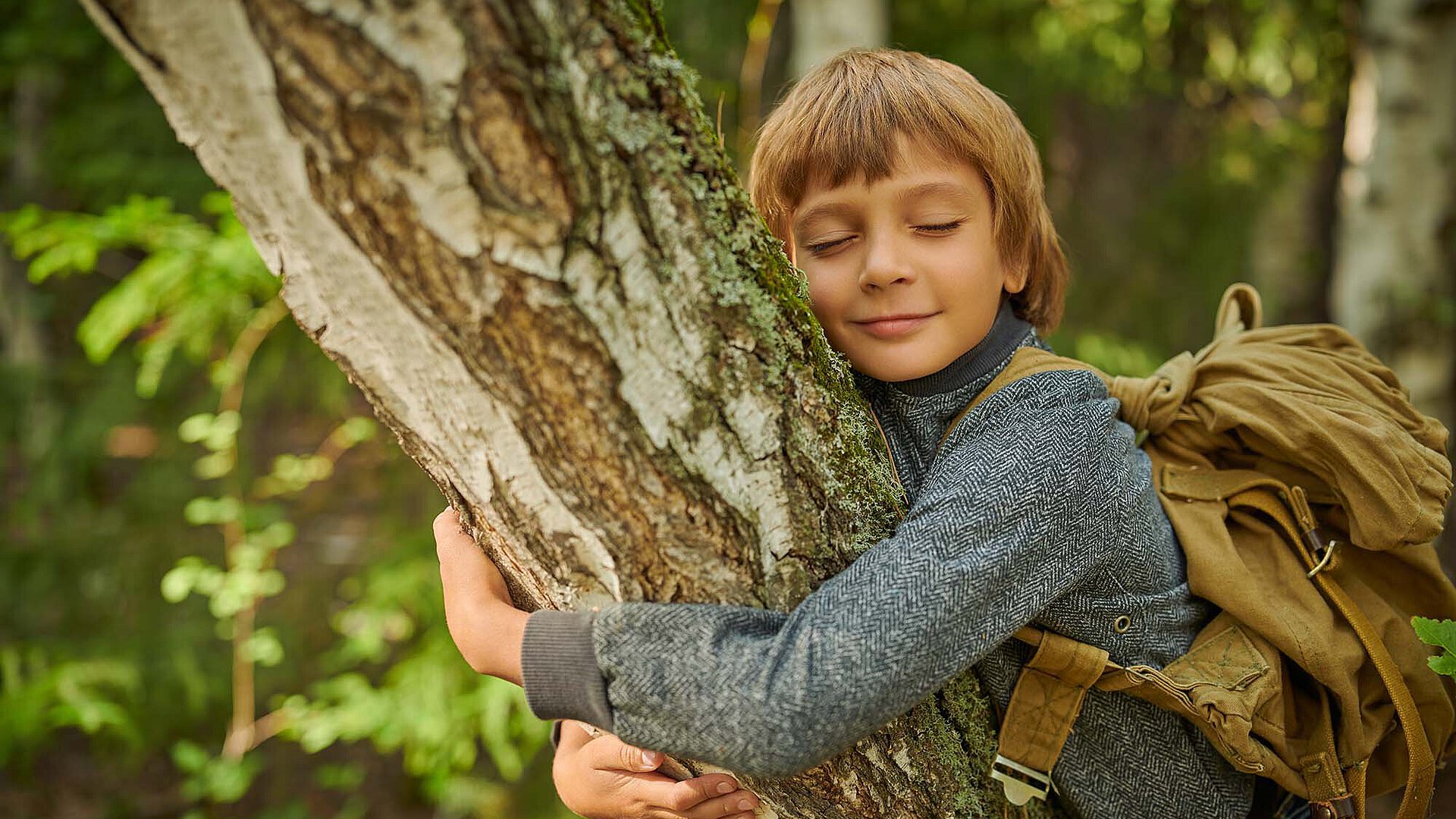 Kind mit Rucksack umarmt im Wald einen Baumstamm, die Augen geschlossen und das Gesicht an der Rinde.