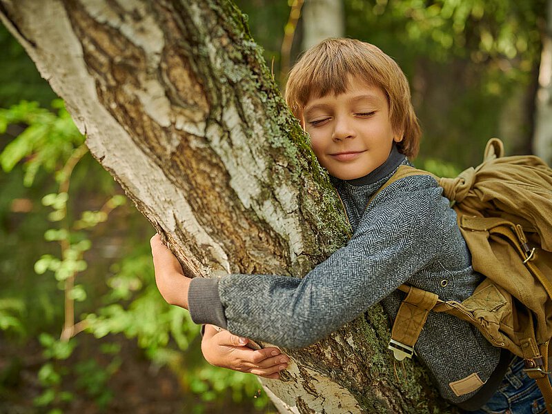 Child with a backpack hugging a tree trunk in the forest, eyes closed and face resting against the bark.