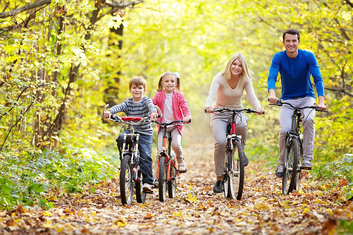 A family of four rides bicycles along a leafy trail in a forest during autumn, with colorful leaves scattered on the ground and trees in the background.