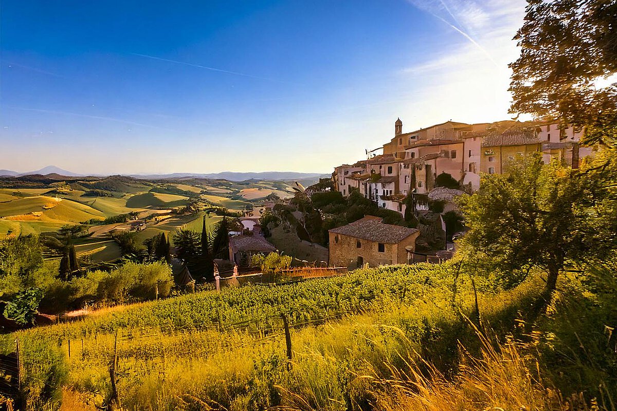 A panoramic view of the Tuscan countryside with rolling hills, vineyards, and a village on a hill, bathed in warm sunset light.