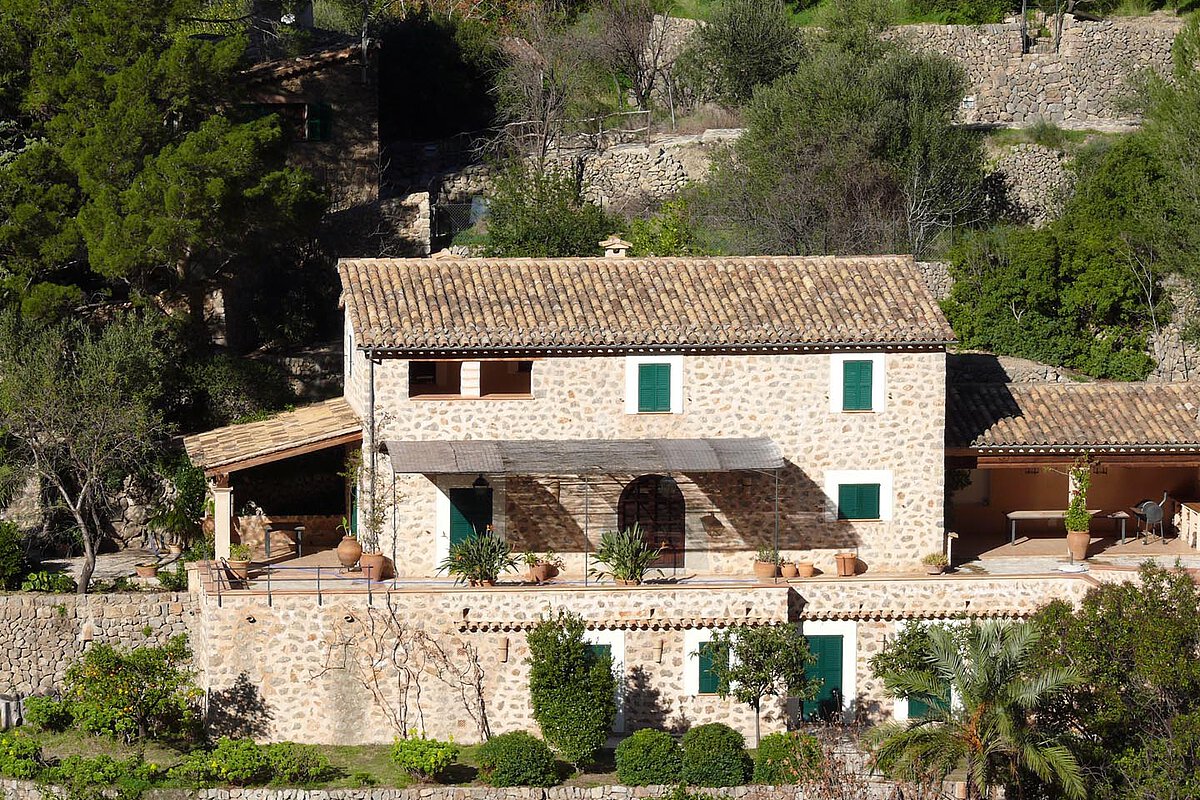 Rustic stone house with a tiled roof and green shutters, surrounded by gardens and trees, set in the tranquil Spanish countryside.