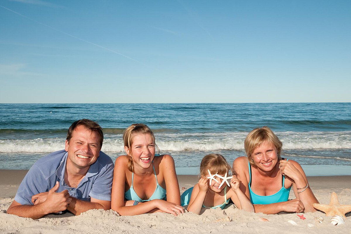 A family of four lying on the sand at the beach, smiling and enjoying the sun, with one child playfully holding a starfish in front of her face and the ocean in the background.
