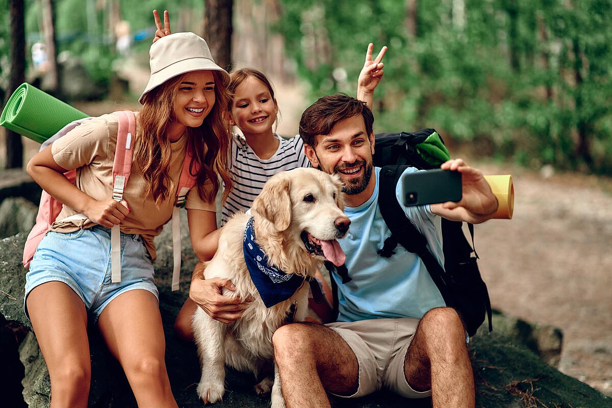 A family poses for a selfie in the woods with their golden retriever. The woman and child are smiling, and the man holds a smartphone, capturing the moment amidst a lush green background.