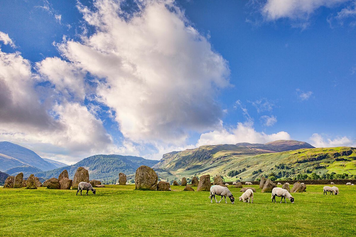 Eine malerische Szene von Schafen, die auf grünem Gras neben großen Steinformationen grasen, mit sanften Hügeln und einem blauen Himmel, der mit Wolken gesprenkelt ist, im Hintergrund.