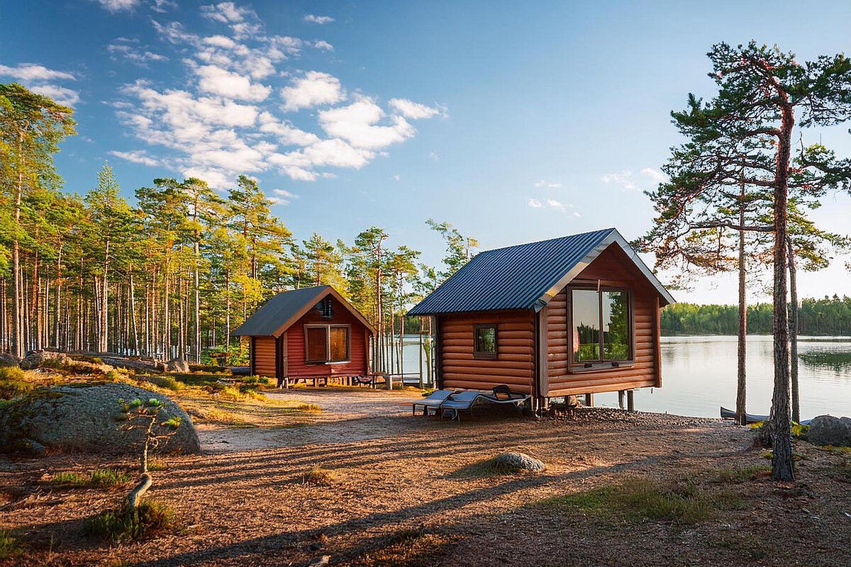 Zwei Holzhütten in Småland, Schweden, die an einem ruhigen See liegen und von hohen Bäumen umgeben sind, während das Sonnenlicht Schatten auf den Boden wirft.