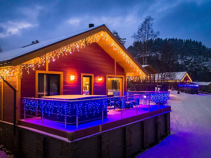 Ein Holzhaus im Schnee mit warmem Licht und Weihnachtsbeleuchtung in den Bergen.