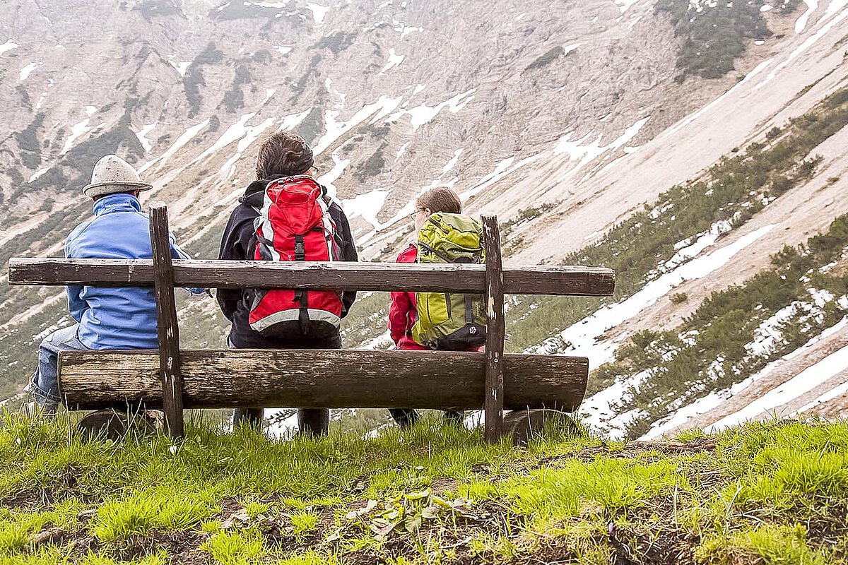 Eine Familie mit drei Personen sitzt auf einer Holzbank und blickt auf eine bergige Landschaft mit Schnee und Grün. Sie sind in Outdoor-Bekleidung gekleidet und genießen die Aussicht, während sie einen Moment der Entspannung in der ruhigen Umgebung genießen.