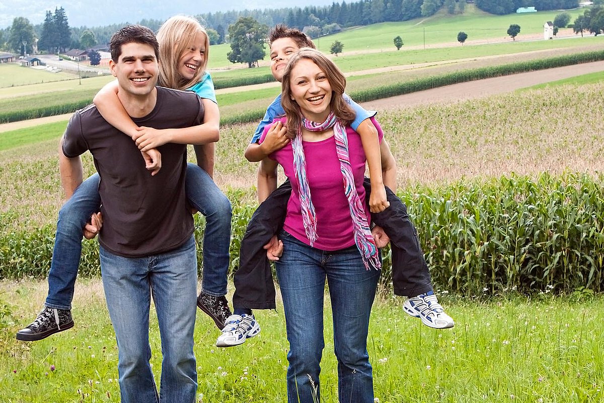 A family with two adults and two children playing outdoors in a green field, with the children on the adults' backs, against a backdrop of hills and farmland.