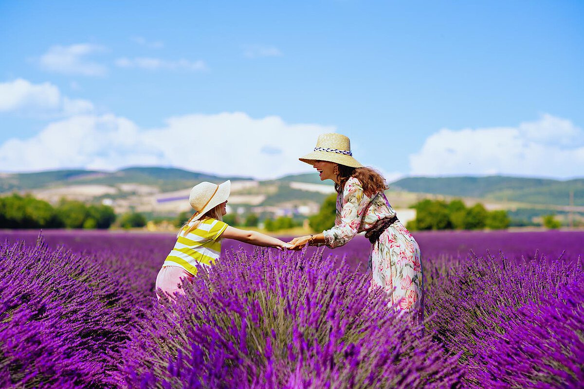 Eine Mutter und Tochter, die Händchen haltend in einem hellen Lavendelfeld stehen, umgeben von lebhaften lila Blumen unter einem sonnigen Himmel, beide mit Sonnenhüten.