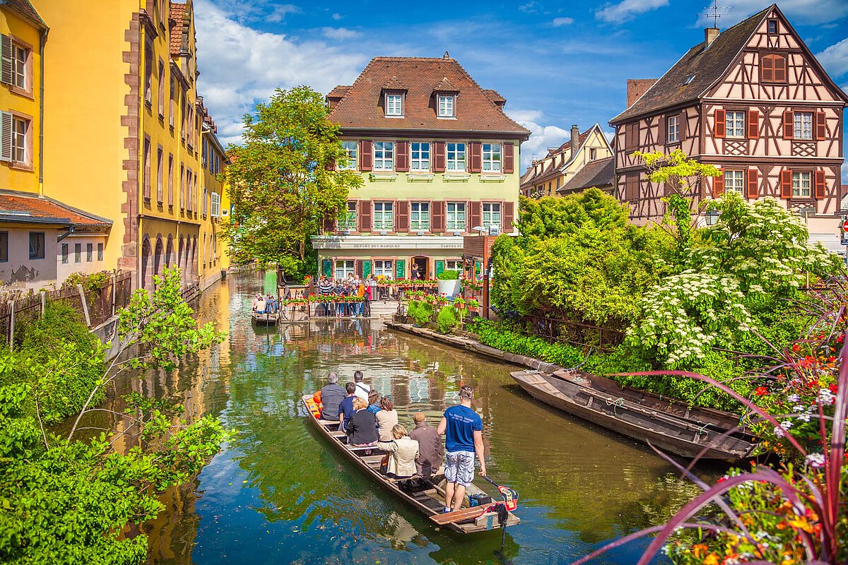 Colorful houses along a canal in Colmar, with people on a boat surrounded by greenery.