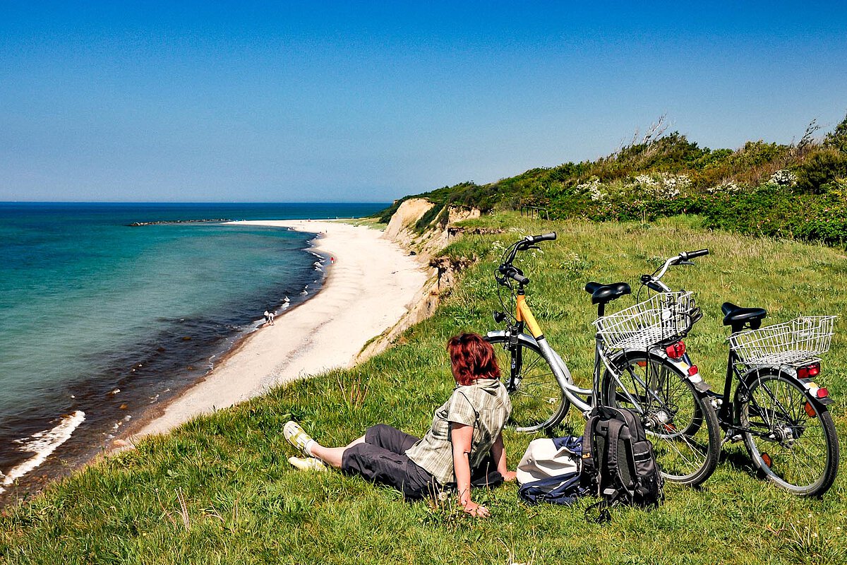 Eine Frau, die sich auf einer grasbewachsenen Klippe in Norfolk entspannt, mit Fahrrädern in der Nähe, die auf einen Sandstrand und das türkisfarbene Meer unter einem klaren blauen Himmel blickt.
