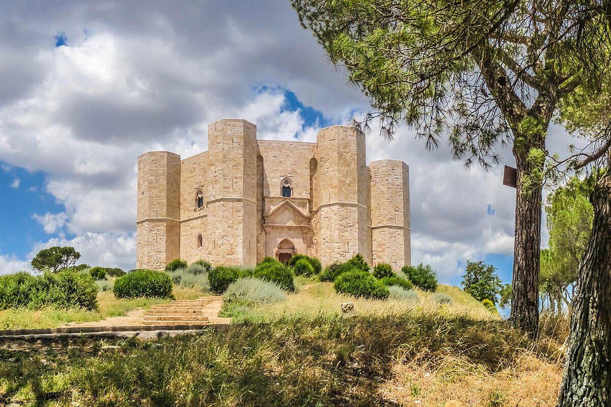A view of Castel del Monte in Salento, showcasing its octagonal stone structure set on a grassy hill, surrounded by trees and a sky filled with clouds.