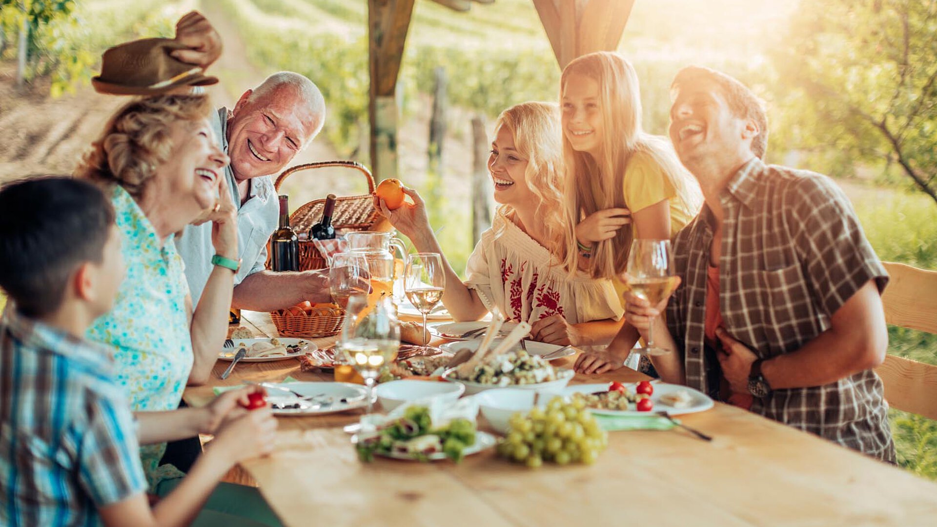 A happy family sitting around a wooden table outdoors, enjoying food and drinks, with laughter and smiles in a vineyard setting illuminated by warm sunlight.