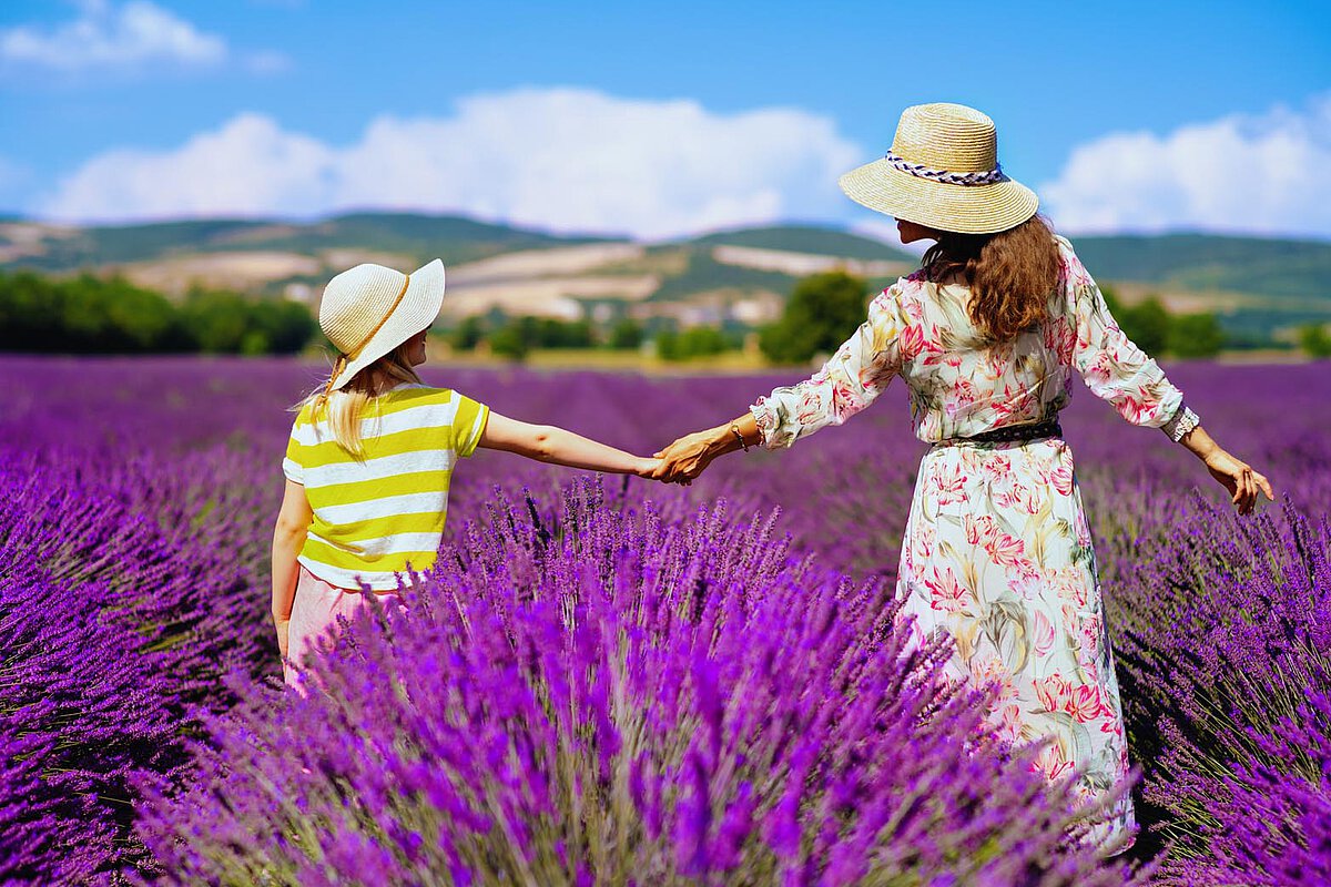 Eine Mutter und Tochter, die Händchen haltend durch ein Lavendelfeld gehen, umgeben von blühenden lila Blumen und einem strahlend blauen Himmel.