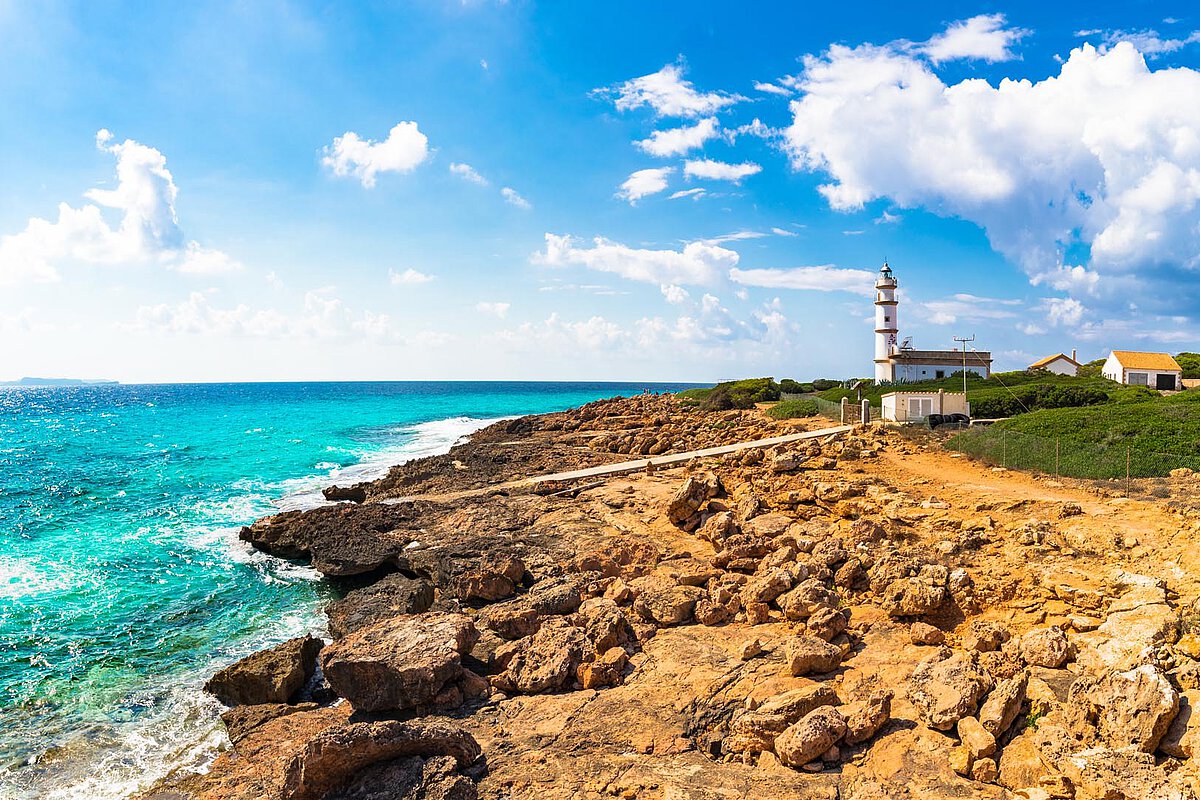 Lighthouse on rocky coastline with turquoise waters in the Canary Islands, under a bright blue sky with scattered clouds, depicting a picturesque seascape.