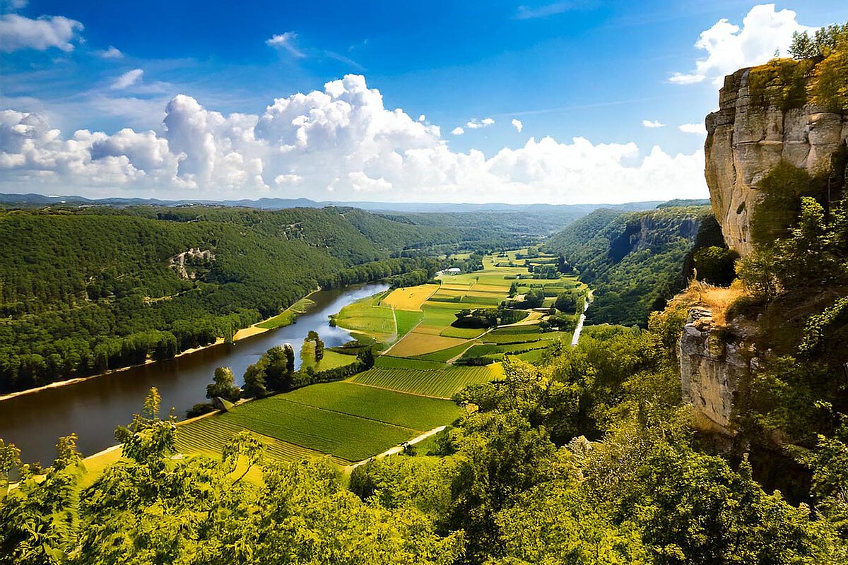 Lush green hills and vineyards along the winding river in the Dordogne Valley, with rocky cliffs and a bright blue sky.