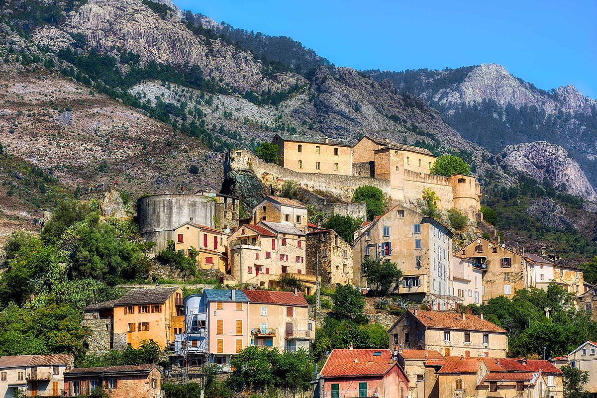 The old town of Corte in Corsica with its citadel perched on a rocky hill, surrounded by colorful houses and mountain scenery under a clear blue sky.