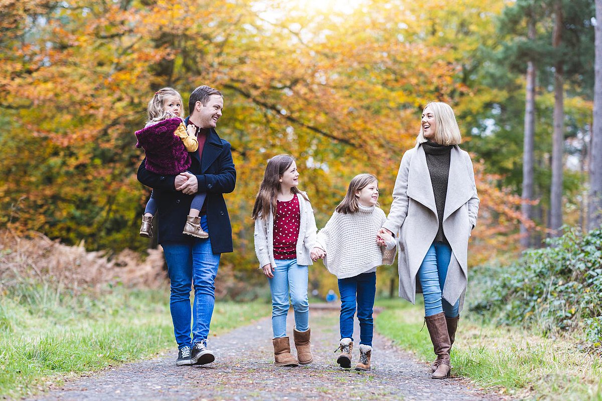 Eine Familie mit fünf Personen genießt einen Spaziergang auf einem Kiesweg im Wald während des Herbstes. Die Eltern gehen Hand in Hand mit ihren drei Kindern, von denen eines auf der Schulter des Vaters sitzt, umgeben von lebhaften Herbstblättern.