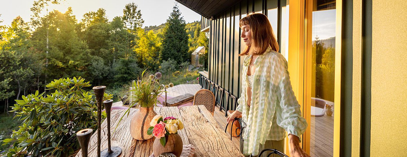 Eine Frau steht auf einem Balkon mit einem Holztisch, der mit Blumen dekoriert ist. Die Szene ist von Grün umgeben und in sanftes Sonnenlicht getaucht, was eine warme und einladende Stimmung erzeugt.