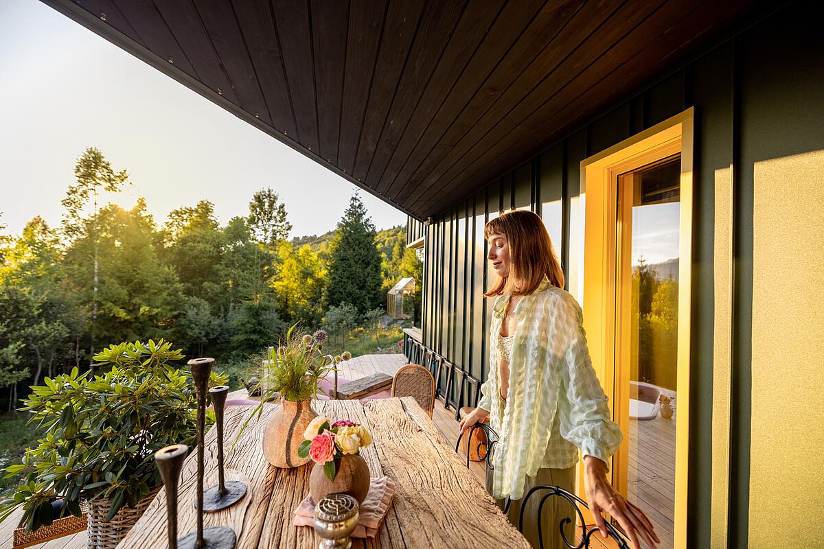 A woman standing on a balcony with a wooden table decorated with flowers. The scene is surrounded by greenery and bathed in soft sunlight, creating a warm and inviting ambiance.
