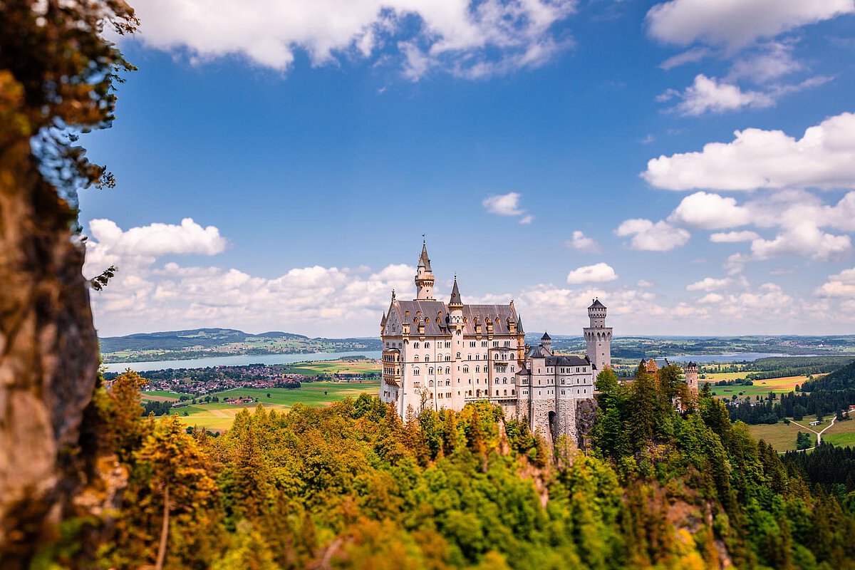 Schloss Neuschwanstein auf einem felsigen Hang, umgeben von Bäumen und Hügeln, unter einem bewölkten Himmel.