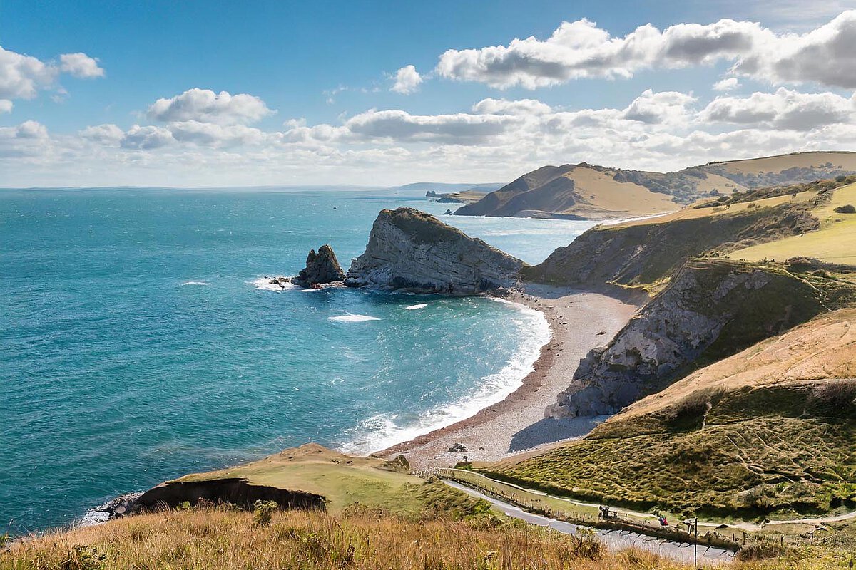 Panoramablick auf die Küste von Dorset mit rauen Klippen, einem kieseligen Strand und sanften Hügeln, während das Meer unter einem teilweise bewölkten Himmel schimmert.
