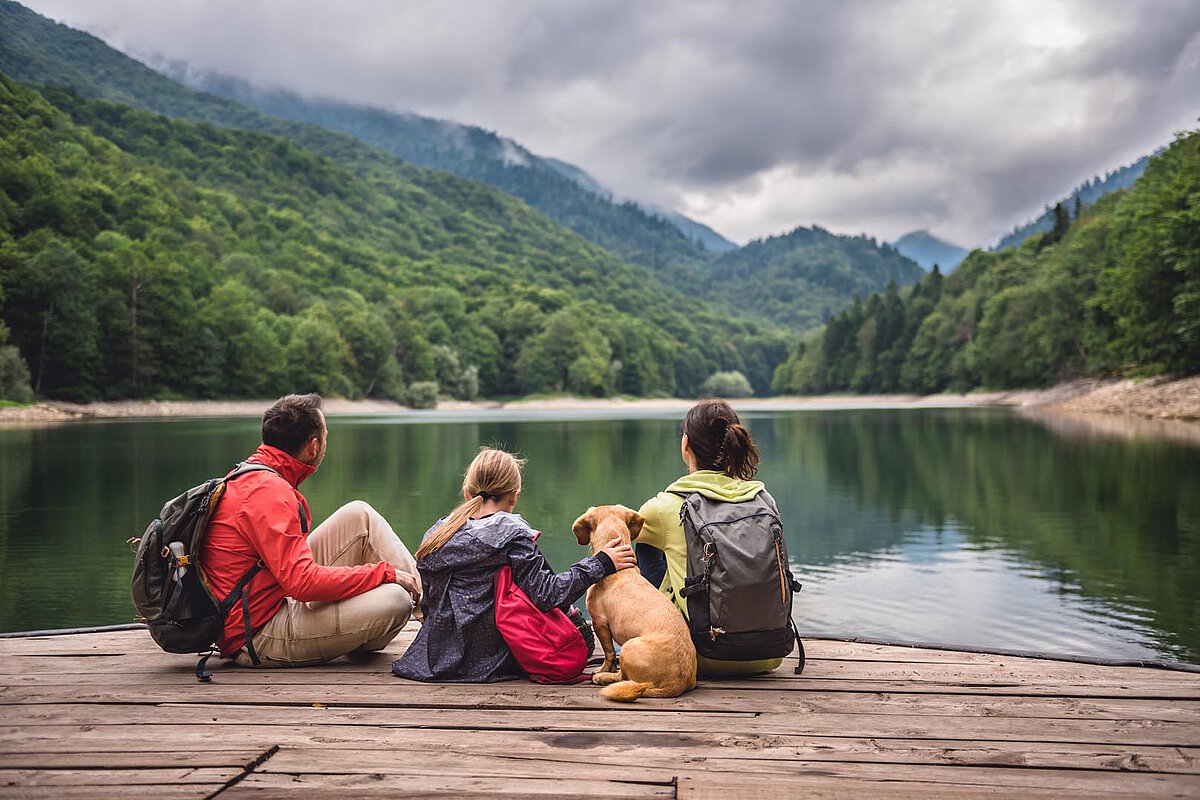 Eine Familie sitzt auf einem Steg am See, mit ihrem Hund neben ihnen. Sie sind von Bergen und Grün umgeben und genießen die entspannte Atmosphäre an einem bewölkten Tag.