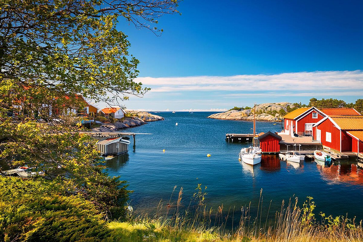 Ein charmanter Hafen an Schwedens Westküste mit roten Holzhäusern und festgemachten Booten, die sich im ruhigen Wasser spiegeln, umgeben von felsigen Vorsprüngen und Grün.
