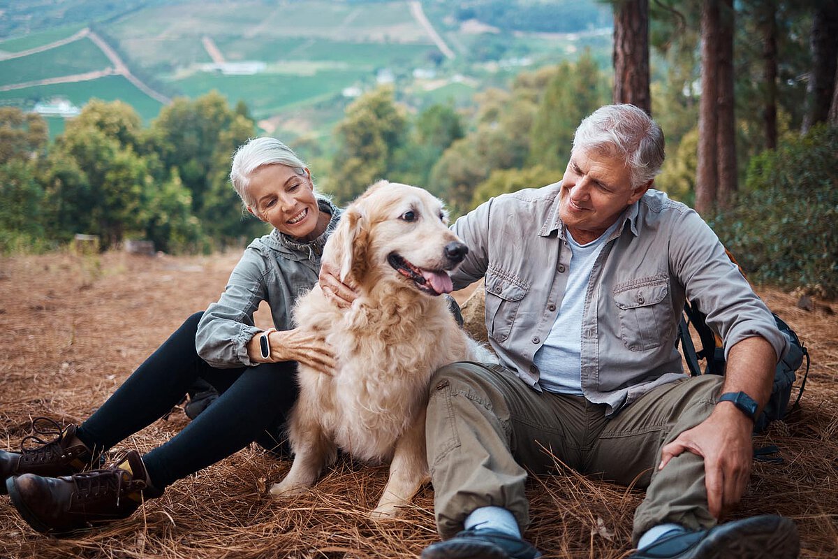 A couple sitting on the ground with their golden retriever in a wooded area. They are smiling and engaging with the dog, with a scenic view of hills and trees in the background.