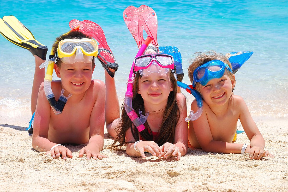 Three children lay on the sand at the beach, wearing snorkeling masks and fins. They smile at the camera, with the vibrant blue water in the background, ready for a fun day of swimming and exploration.