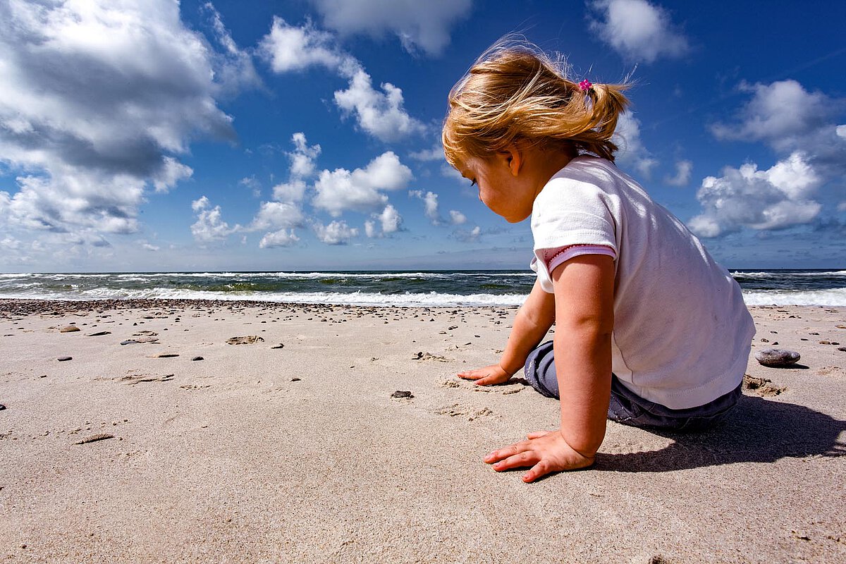 Ein kleines Kind sitzt am sandigen Strand, dem Ozean zugewandt. Das Kind trägt ein leichtes Shirt und Jeans, mit zurückgebundenem Haar, und genießt einen sonnigen Tag am Wasser.