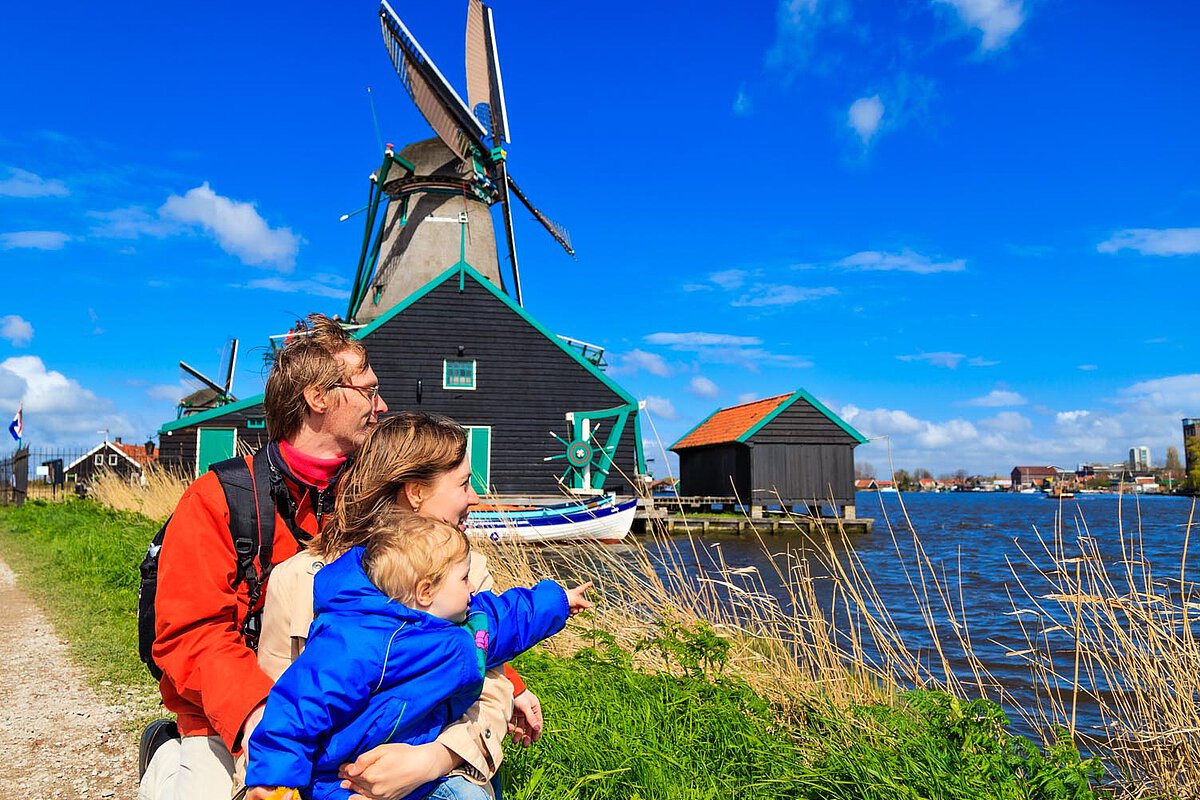 A family sits by the water, with a windmill and traditional houses in the background. The parents are looking out, and the mother is pointing while the child sits on the father's lap, dressed in a blue jacket.